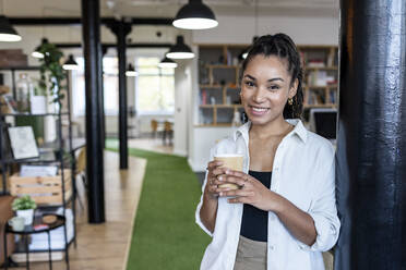 Smiling young businesswoman with disposable coffee cup leaning on column in office - WPEF07079