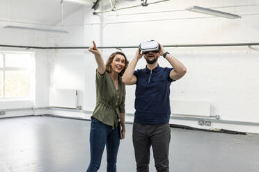 Happy young businessman wearing virtual reality simulator and colleague pointing in empty space - WPEF07100