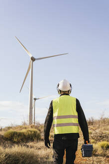 Techniker mit Werkzeugkasten vor einer Windkraftanlage an einem sonnigen Tag - MGRF00857