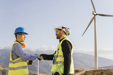Engineer shaking hands with technician in front of wind turbines - MGRF00879