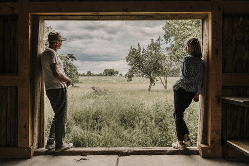Man and woman looking at field leaning on doorway - MJRF00863