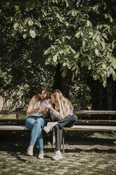 Frau teilt Smartphone mit Freund auf Bank unter Baum sitzend - MJRF00868