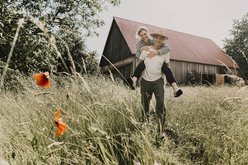 Happy man piggybacking woman in front of barn - MJRF00881