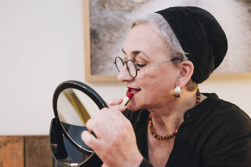 Senior woman applying red lipstick looking in mirror at home - NGF00789