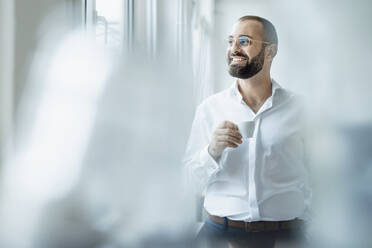 Happy businessman holding cup of coffee at home office - JOSEF15955