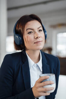 Kontemplative Geschäftsfrau, die mit einer Kaffeetasse im Büro über Kopfhörer Musik hört - JOSEF15991