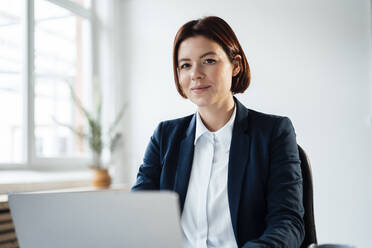 Smiling businesswoman sitting with laptop in office - JOSEF15993