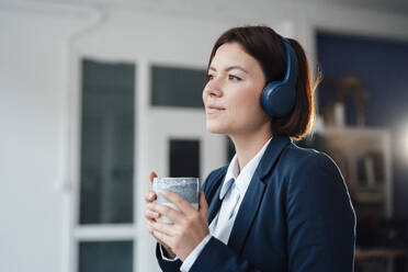 Nachdenkliche Geschäftsfrau, die mit einer Kaffeetasse im Büro über Kopfhörer Musik hört - JOSEF16002