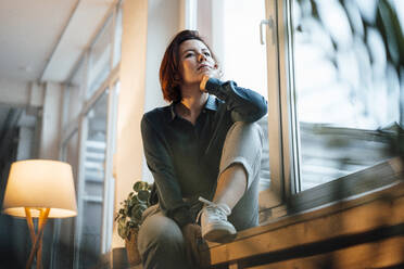Nachdenkliche Geschäftsfrau auf der Fensterbank am Arbeitsplatz sitzend - JOSEF16005