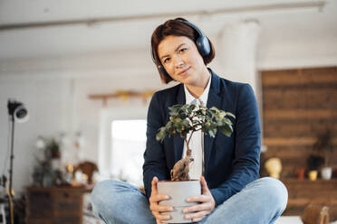 Smiling businesswoman with potted plant listening to music through headphones in office - JOSEF16010