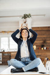 Lächelnde junge Geschäftsfrau, die im Büro eine Topfpflanze auf dem Kopf trägt - JOSEF16011