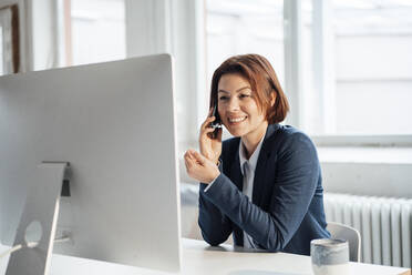 Glückliche Geschäftsfrau, die am Arbeitsplatz vor dem Schreibtisch mit ihrem Smartphone telefoniert - JOSEF16013