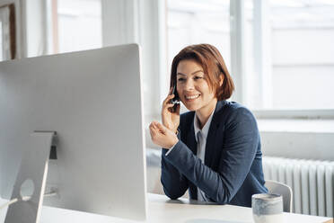 Happy businesswoman talking on smart phone in front of desktop at workplace - JOSEF16013