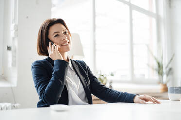 Glückliche Geschäftsfrau, die am Schreibtisch im Büro über ihr Mobiltelefon spricht - JOSEF16015