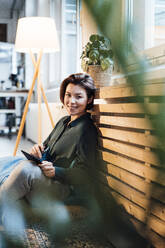 Happy young businesswoman sitting by heating radiator in office - JOSEF16050