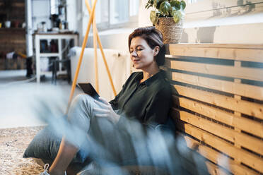 Smiling young businesswoman using tablet computer sitting radiator in office - JOSEF16052