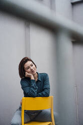 Thoughtful businesswoman sitting on yellow chair in front of gray wall - JOSEF16066