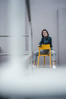 Junge Geschäftsfrau mit Hand am Kinn vor einer grauen Wand sitzend - JOSEF16068