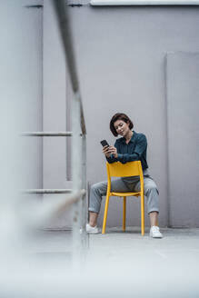 Junge Geschäftsfrau, die ein Smartphone benutzt und auf einem Stuhl vor einer Wand sitzt - JOSEF16069