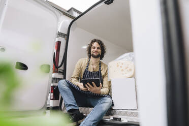 Lächelnder junger Geschäftsmann sitzt mit Tablet-PC und Käse im Lieferwagen - JOSEF16140