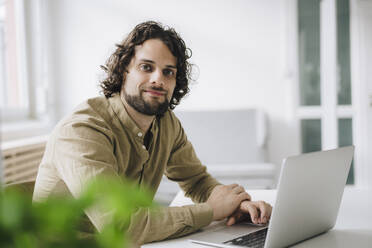 Smiling young businessman sitting with laptop at desk in office - JOSEF16152