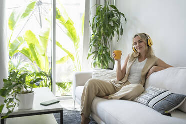 Lächelnde Frau mit drahtlosen Kopfhörern und Kaffeetasse auf dem Sofa zu Hause - SVKF01097