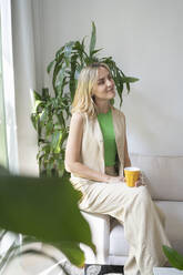 Thoughtful woman sitting with coffee cup on sofa at home - SVKF01104