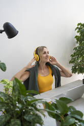 Smiling freelancer wearing wireless headphones sitting at desk in home office - SVKF01135