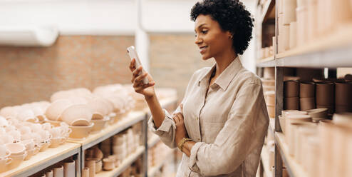 Female entrepreneur reading a text message on her smartphone. Happy businesswoman managing a store with handmade ceramic products. Creative female ceramist running a successful small business. - JLPSF28993