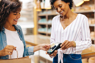 Satisfied female customer making a contactless credit card payment in a ceramic store. Cheerful young woman smiling happily while shopping from a local female-owned small business. - JLPSF28994