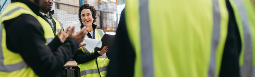 Ältere Frau, die während einer Besprechung mit ihren Kollegen in einem Auslieferungslager fröhlich lächelt. Gruppe von multikulturellen Mitarbeitern, die in einem großen Logistikzentrum zusammenarbeiten. - JLPSF29000