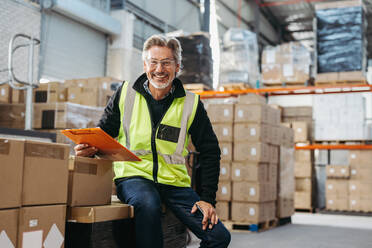 Happy senior man smiling at the camera while holding a clipboard in a distribution warehouse. Cheerful logistics manager wearing a reflective jacket in a large fulfillment centre. - JLPSF29010