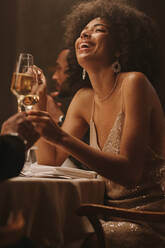 African woman toasting drink with a friend at dinner party. Cheerful female celebrating with drinks at a gala dinner event. - JLPSF29030