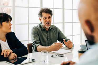 Senior business man discussing with his team, giving solutions and ideas. He talks to his colleagues while they sit on a conference table in an office. - JLPSF29041