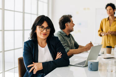 Professionelle Frau, die in einem Sitzungssaal sitzt, mit ihren Kollegen im Hintergrund. Glückliche Geschäftsfrau, die in die Kamera schaut. Frau, die mit einem kreativen Team in einem Büro arbeitet. - JLPSF29065