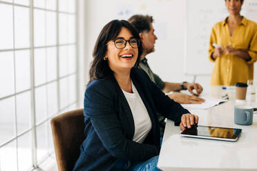 Successful business woman sitting in a boardroom with her colleagues. Business woman smiling at the camera in an office. Professional woman having a meeting with team. - JLPSF29067