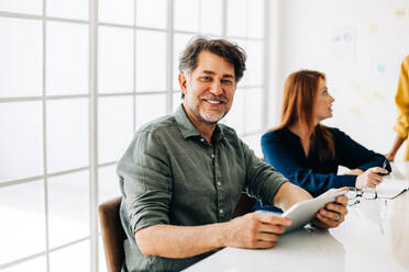 Senior Business Professional hält ein Tablet in einem Sitzungssaal. Glücklicher Geschäftsmann sitzt in einer Besprechung mit seinen Kollegen. Reifer Geschäftsmann arbeitet mit seinem Team in einem Büro. - JLPSF29068