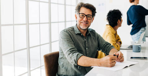 Happy business man looking at the camera while sitting in a boardroom. Senior man attending a meeting in an office. Speech presentation in the background. - JLPSF29073