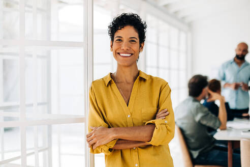Portrait of a black business woman standing in an office with her colleagues in the background. Happy female professional working in a marketing agency. - JLPSF29082