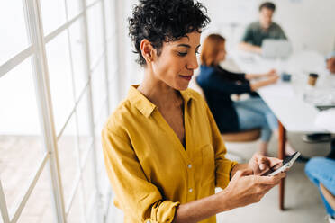 Geschäftsfrau, die bei der Arbeit eine Textnachricht auf einem Mobiltelefon tippt. Geschäftsfrau, die während einer Besprechung eine Textnachricht schreibt. Kollegen, die im Hintergrund eine Besprechung abhalten. - JLPSF29089