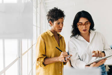 Two business women having a discussion, they're standing in an office and using a tablet. Professional women making a to do list as part of their project planning. - JLPSF29090