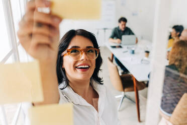 Geschäftsfrau beim Brainstorming mit Haftnotizen in einem Büro. Kreative Geschäftsfrau, die ihre Ideen an eine Glaswand schreibt. Berufstätige Frau, die in einer Marketing-Agentur an einer Geschäftsstrategie arbeitet. - JLPSF29092