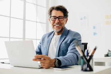 Älterer Softwareentwickler, der an einem Laptop in einem Büro arbeitet. Reifer Geschäftsmann, der in die Kamera schaut, während er an seinem Schreibtisch sitzt. Glücklicher Geschäftsmann, der im technischen Bereich arbeitet. - JLPSF29106