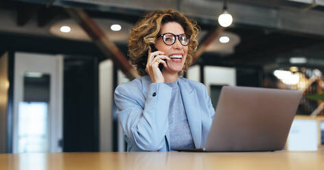 Business woman talking on a phone call in a coworking office. Happy female professional calling her associates on a mobile phone. Professional business woman sitting with a laptop. - JLPSF29125
