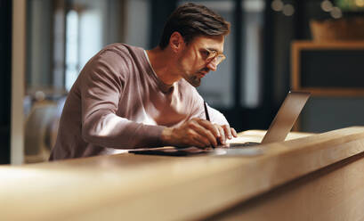 Focused graphic designer drawing on a graphics tablet in a cafe. Business man working on a design with a laptop. Male design professional working remotely. - JLPSF29249