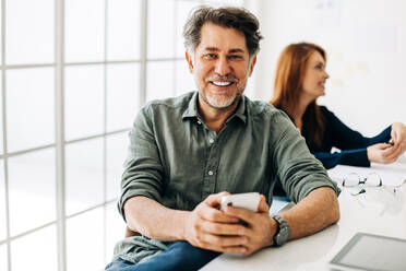 Business man smiling at the camera while sitting in a boardroom. Senior business man using a smart phone in a meeting. Experienced business professional working in a creative office. - JLPSF29297