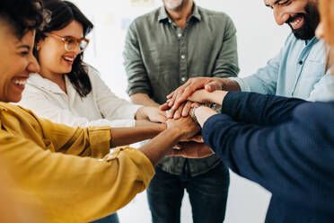 Happy business team stacking their hands together in a huddle. Group of diverse business people having a meeting in an office. Team unity and motivation at work. - JLPSF29304