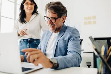 Älterer Geschäftsmann, der in einem Büro an einem Laptop arbeitet. Reifer Softwareentwickler, der mit seinem Kollegen spricht, während er an einem Projekt arbeitet. Glücklicher Geschäftsmann, der in der Technik arbeitet. - JLPSF29313