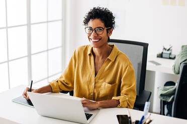 Grafikdesignerin bei der Arbeit am Schreibtisch in einem Büro. Frau lächelt in die Kamera, während sie mit einem Grafiktablett entwirft. Geschäftsfrau sitzt mit einem Laptop in einem Büro. - JLPSF29323