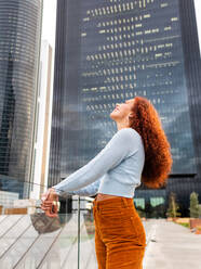 Cheerful young female millennial with long curly red hair and lips in sweater leaning on glass fence and looking up near contemporary skyscraper in city - ADSF42698